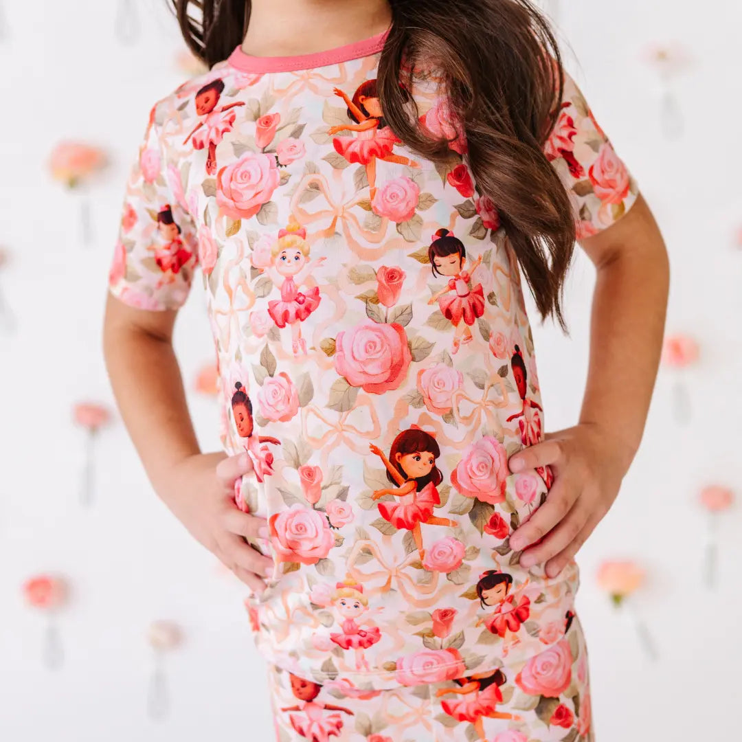 Close-up of a child in a short-sleeve pajama top printed with pink roses, ribbons, and little ballerinas, hands on hips.